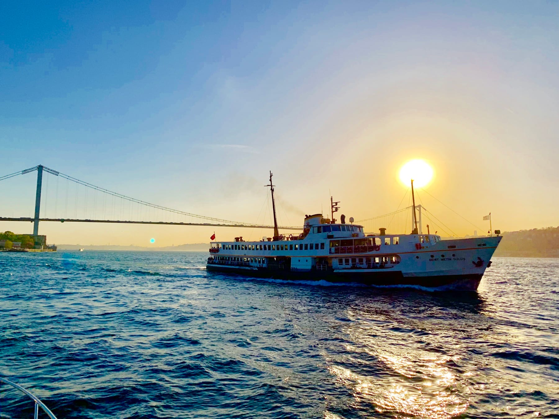 A classic Istanbul ferry crossing the Bosphorus at sunset, the sun low over the water beside the Bosphorus Bridge