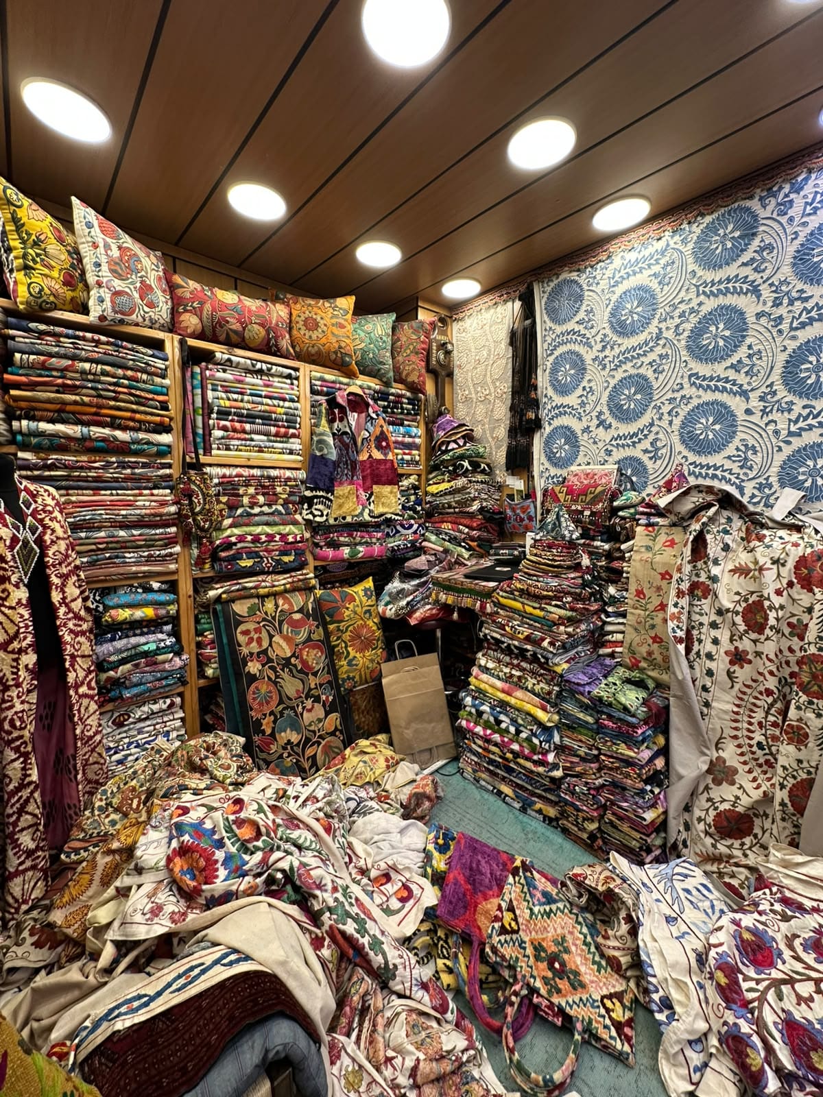 A textile shop in Istanbul’s Grand Bazaar stacked with hand-embroidered suzani, a large blue and white suzani panel hanging on the back wall