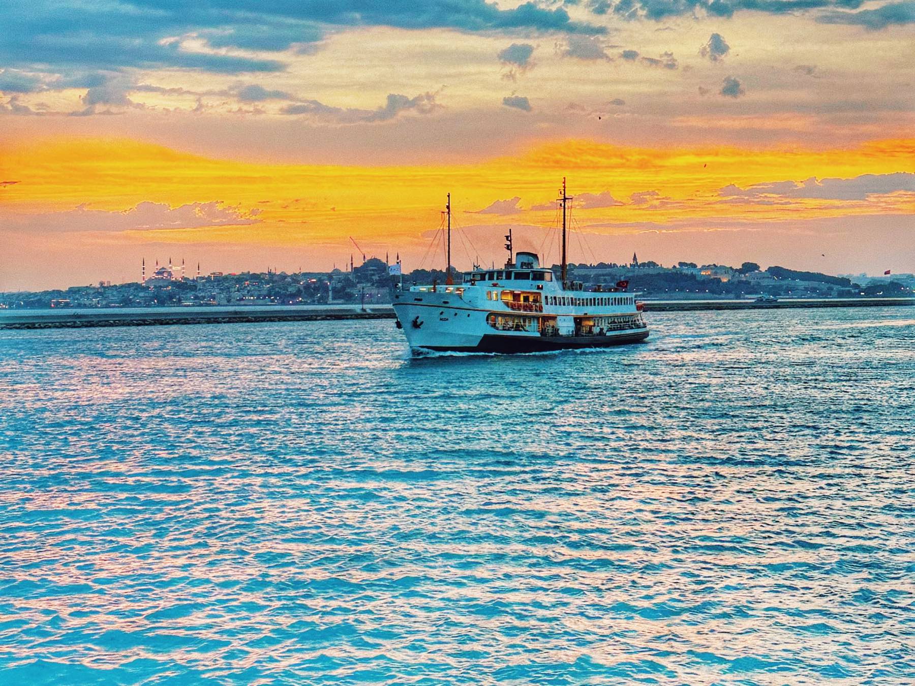 An Istanbul ferry crossing the water in golden evening light, the domes and minarets of the Old City rising on the shore behind it