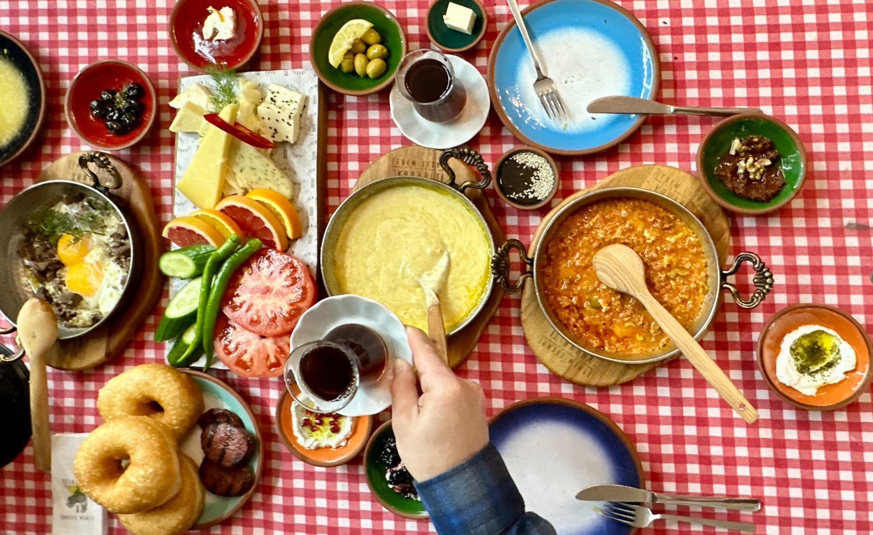 An overhead view of a generous Turkish breakfast spread, with a hand reaching for a glass of tea