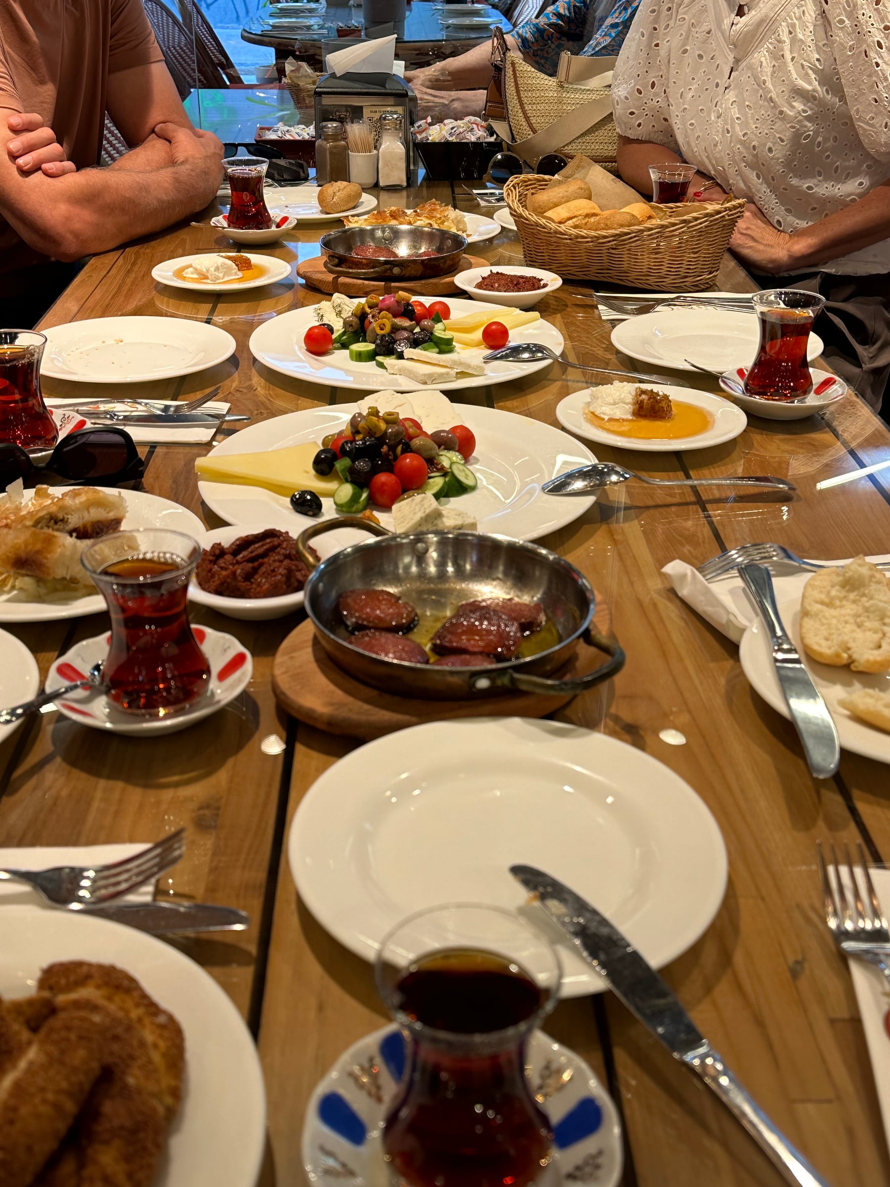 A long wooden table laid for a Turkish breakfast, with plates of cheeses, olives, tomatoes, honey and bread, and tulip glasses of black tea all down its length