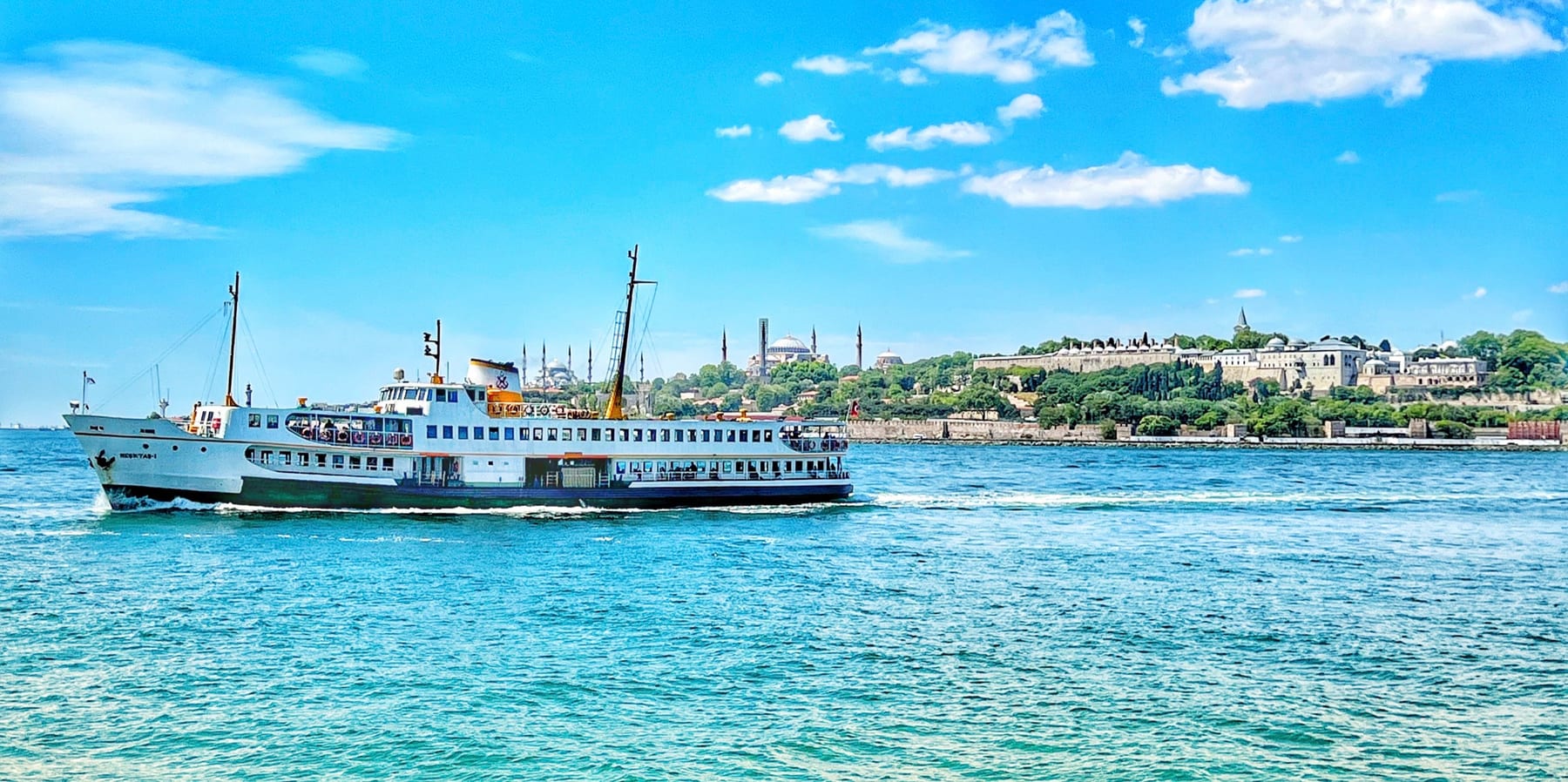 A panoramic view of Istanbul's historic Old City peninsula from the water, a classic ferry crossing in the foreground and the domes and minarets of the Blue Mosque, Hagia Sophia and Topkapi Palace along the skyline