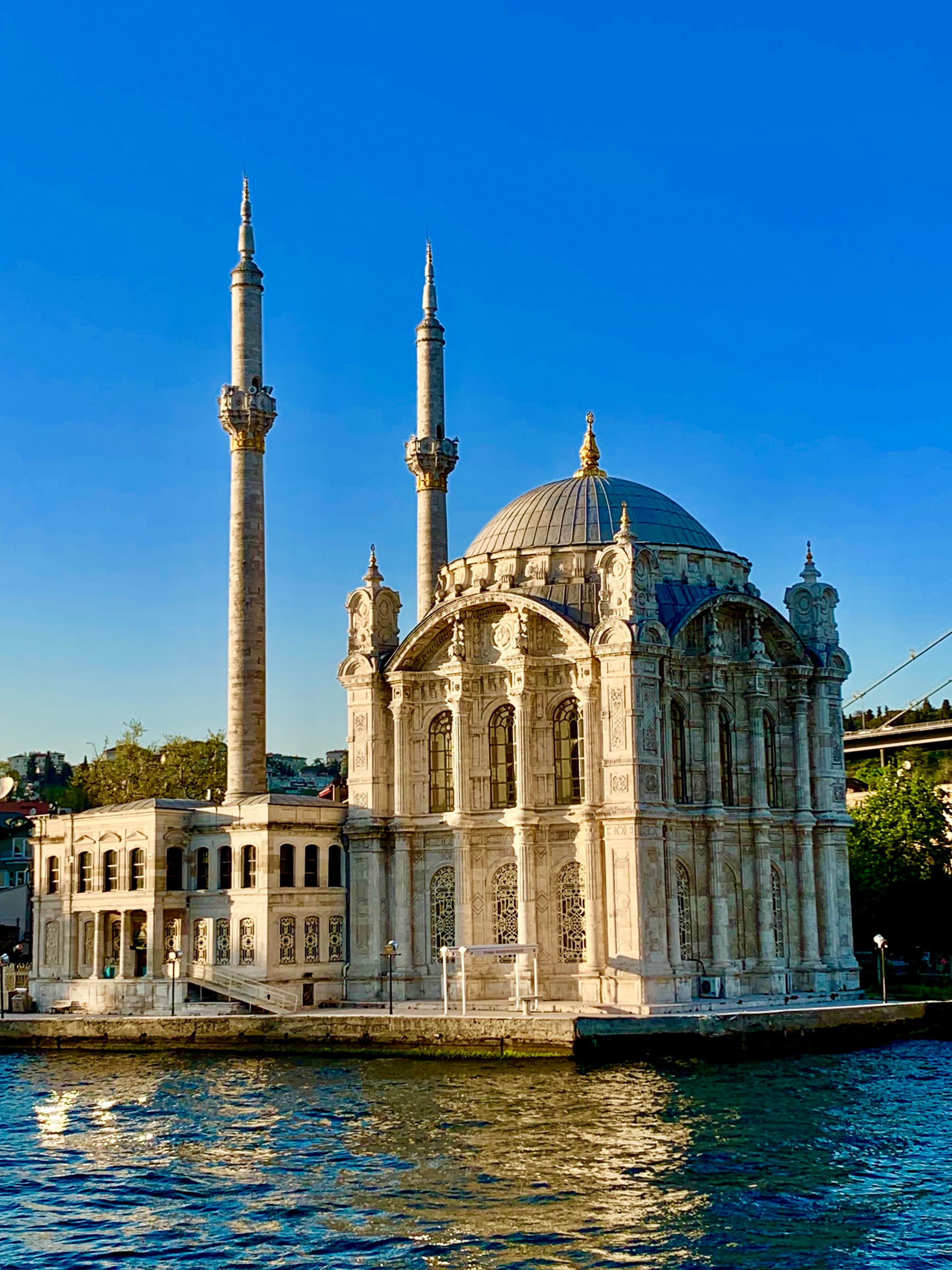 The ornate Ortakoy Mosque on the Bosphorus shoreline, its twin minarets and dome glowing in golden light beneath the Bosphorus Bridge