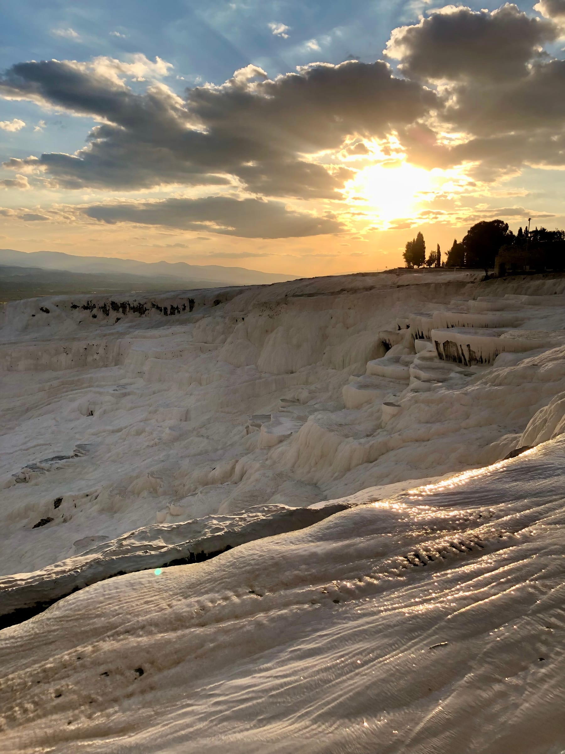 The white travertine terraces of Pamukkale at sunset, thermal water lying in shallow pools across the ridge with the mountains beyond